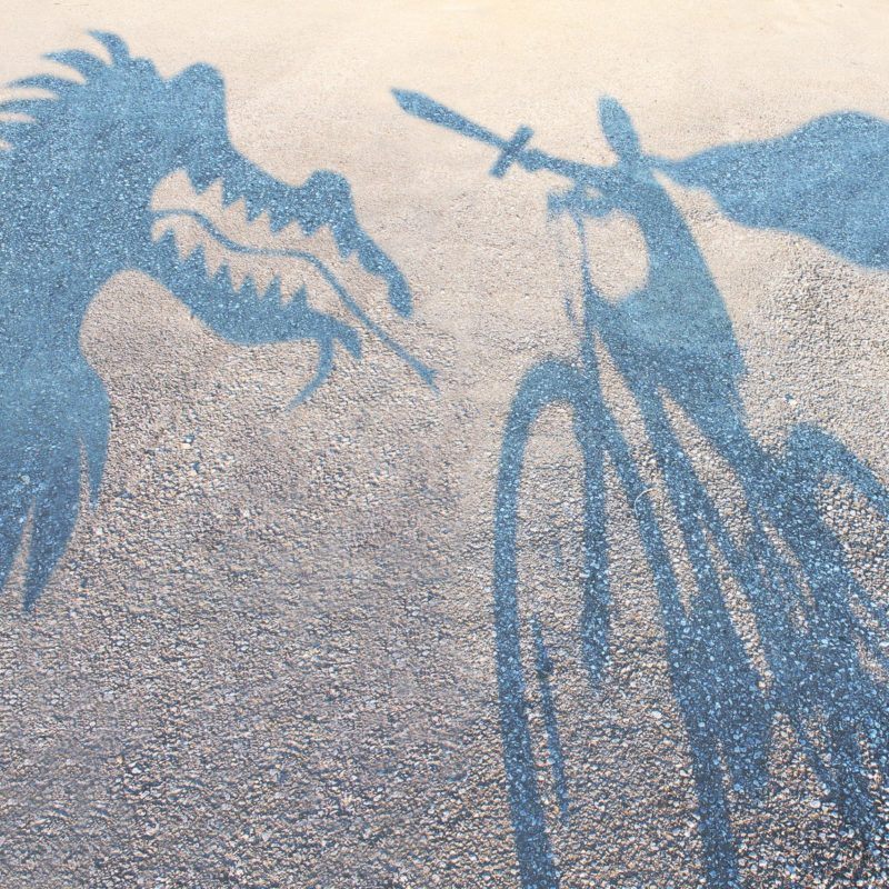 25725299 - children imagination concept with cast shadows on a gravel floor of a superhero child wearing a cape on a bicycle slaying an imaginary dragon