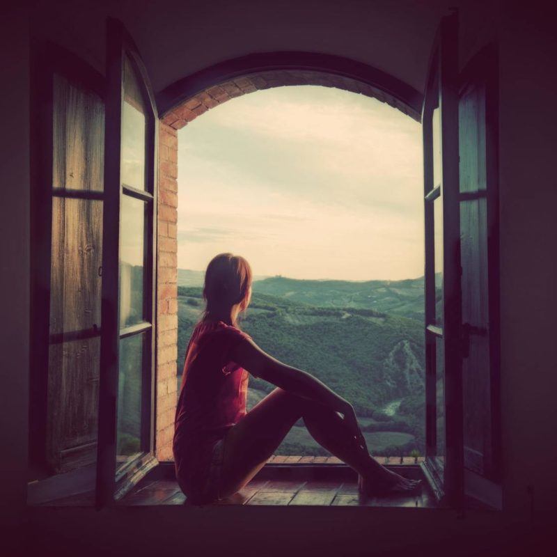 47060552 - young woman sitting in an open old window looking on the landscape of tuscany, italy. conceptual romantic, dreaming, hope, travel.