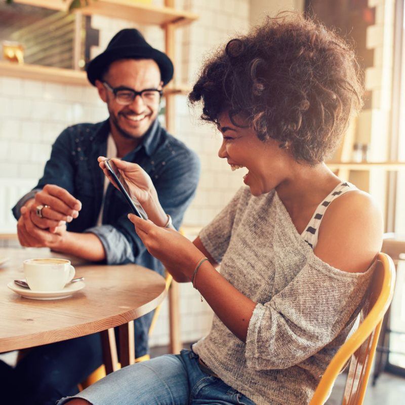 52549157 - portrait of smiling young woman at a cafe table looking at digital tablet with a friend sitting by.