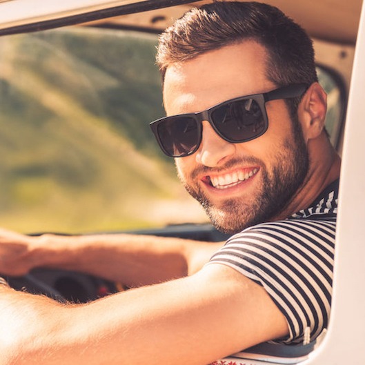 41179559 - enjoying his road trip. cheerful young man smiling at camera and holding hand on steering wheel while sitting inside of his minivan